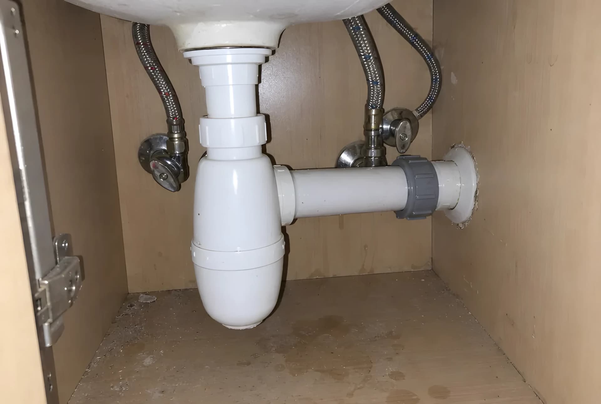Sifón de botella de PVC blanco bajo un lavabo, conectado al desagüe de la pared, con los latiguillos del grifo a los lados