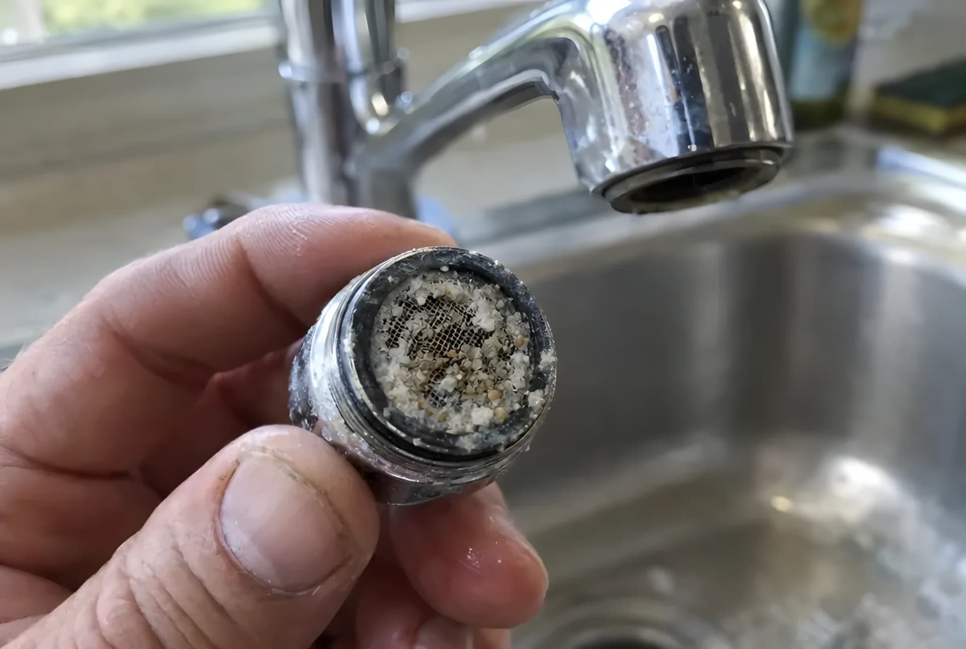 Mano sujetando un aireador de grifo recién desenroscado con la rejilla taponada de cal y arenilla, con el grifo de la cocina al fondo