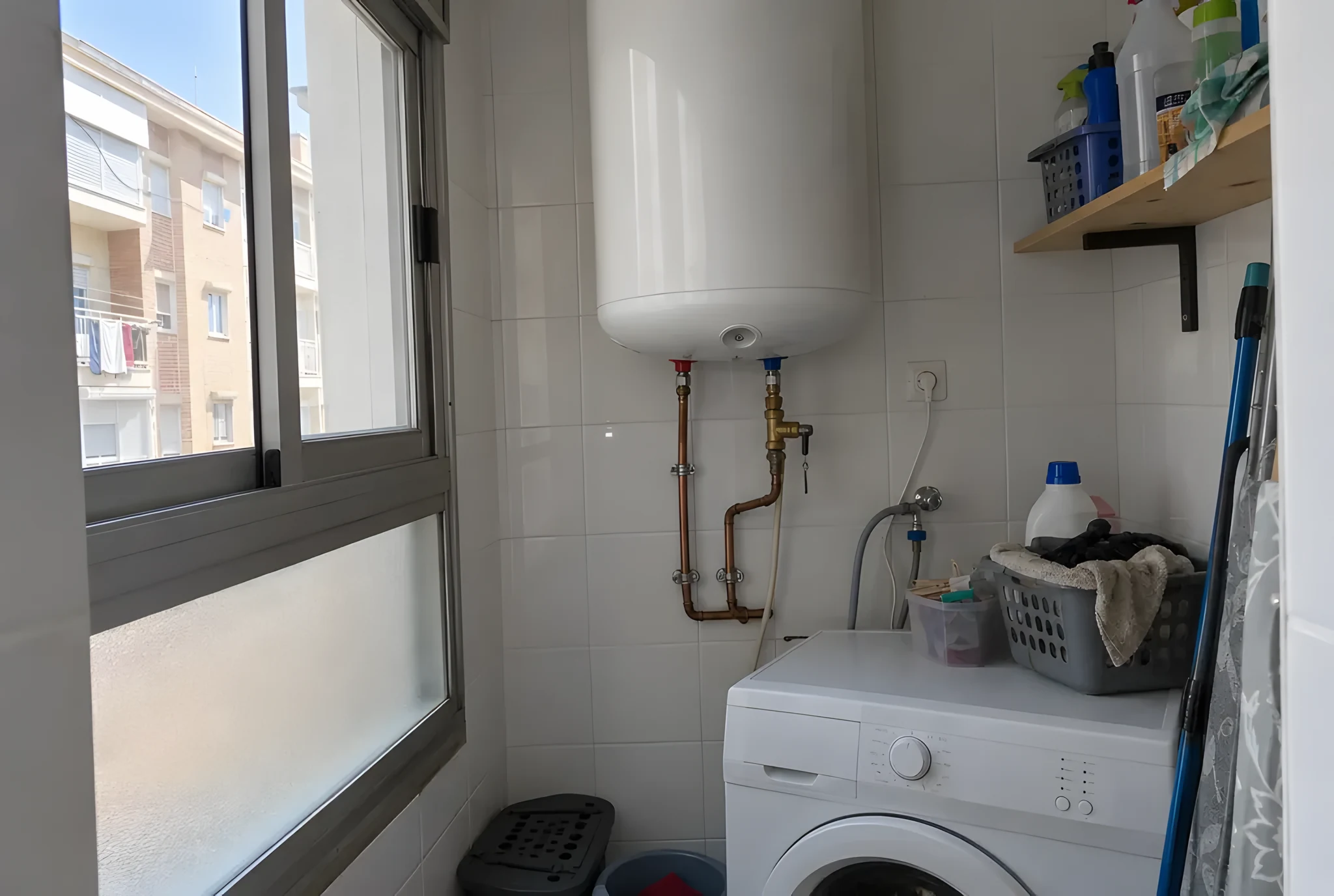 Termo eléctrico vertical blanco colgado en la pared alicatada de una galería, con tuberías de cobre y válvula de latón debajo